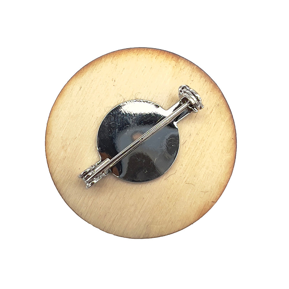 Back view of a circular wooden badge with a metal pin fastening, designed by The Unruly Stitch, showing a top-down perspective.