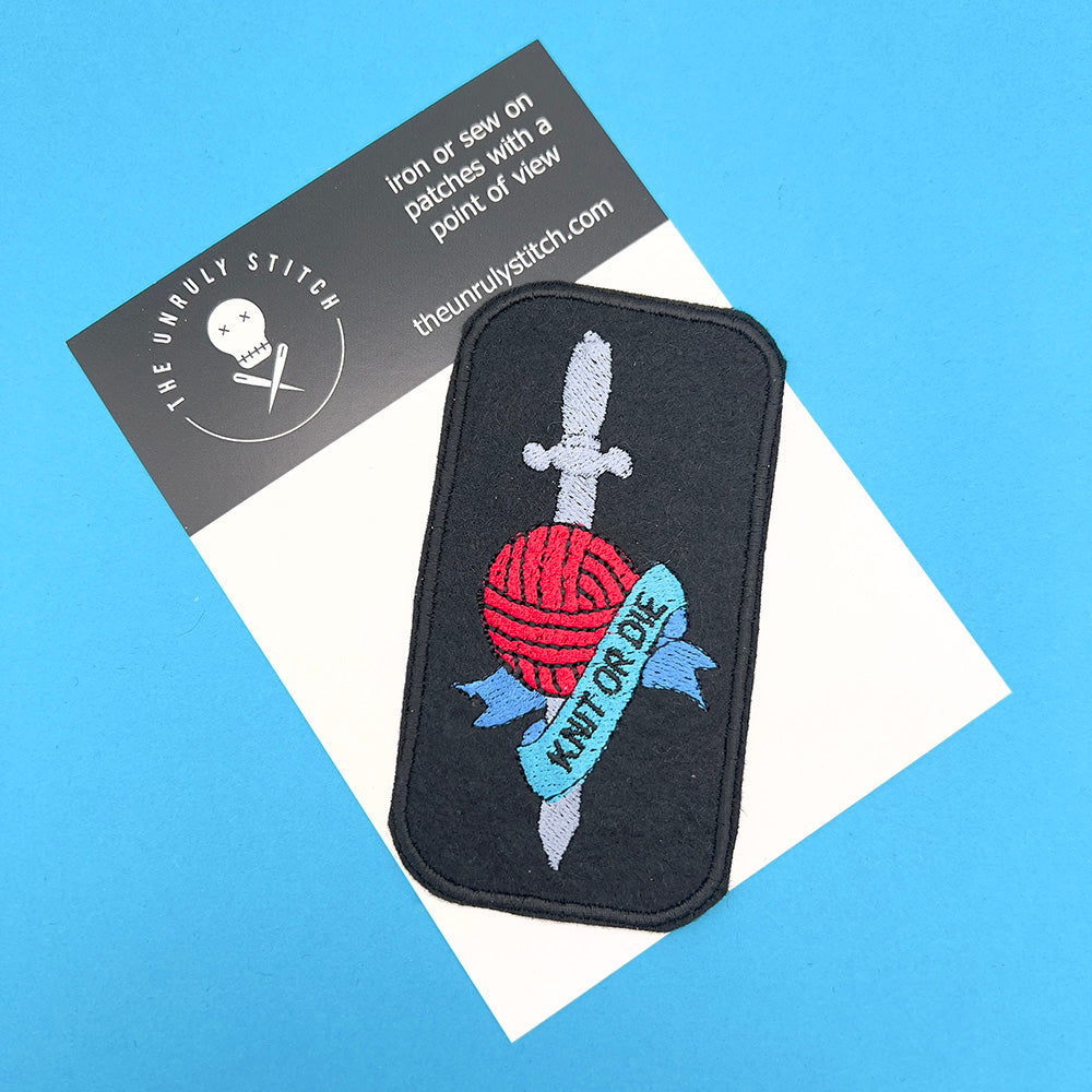 Embroidered felt patch with a red ball of yarn and a silver dagger, wrapped with a blue banner that says "Knit or Die," is attached to a branded card from "The Unruly Stitch."