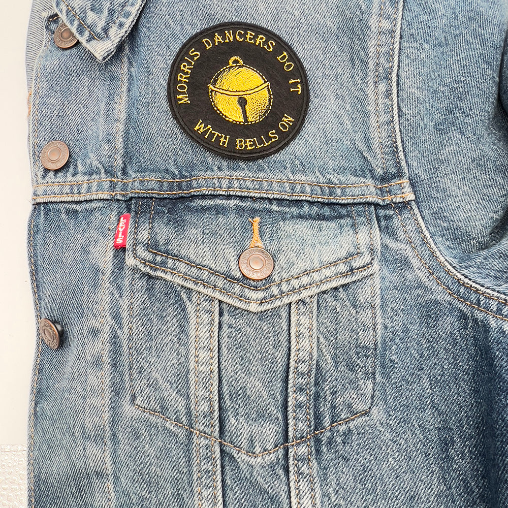 An embroidered felt patch with a yellow bell and the text "Morris Dancers Do It With Bells On" in yellow, sewn above the pocket of a blue denim jacket.