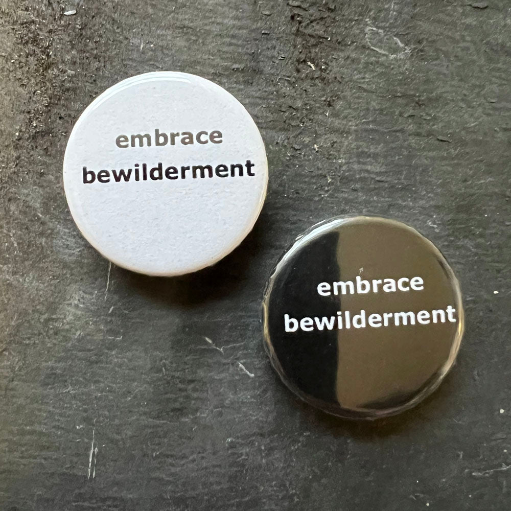 Two circular pin badges side by side on a dark surface. The badge on the left is white with black text that reads "embrace bewilderment." The badge on the right is black with white text that reads "embrace bewilderment.