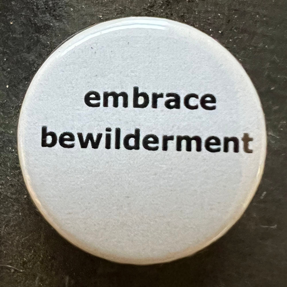 Close-up of a white circular pin badge with black text that reads "embrace bewilderment" against a dark background.