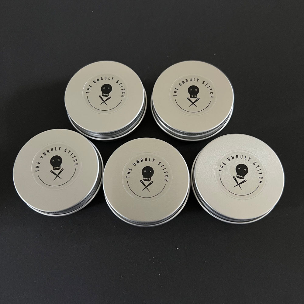 Five round metal tins arranged in a star pattern, each with 'The Unruly Stitch' logo featuring a stylized skull with crossed knitting needles beneath it, on a black background.