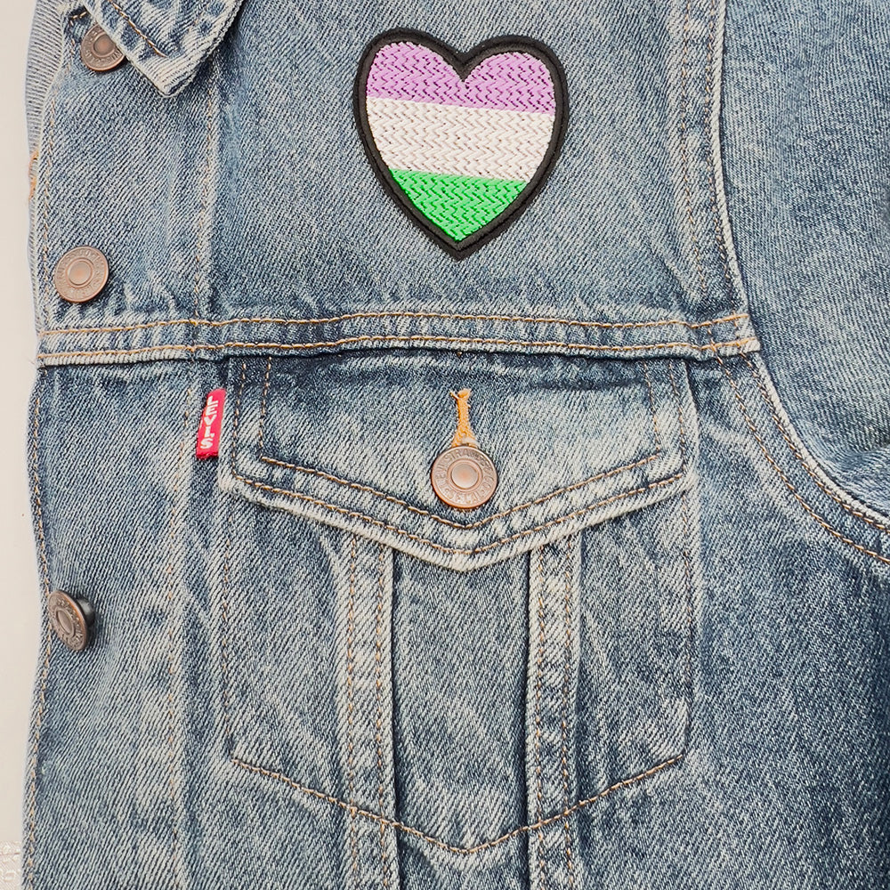 Gender queer pride flag heart-shaped embroidered felt patch attached to the front pocket of a denim jacket. The patch displays horizontal stripes in purple, white, and green.