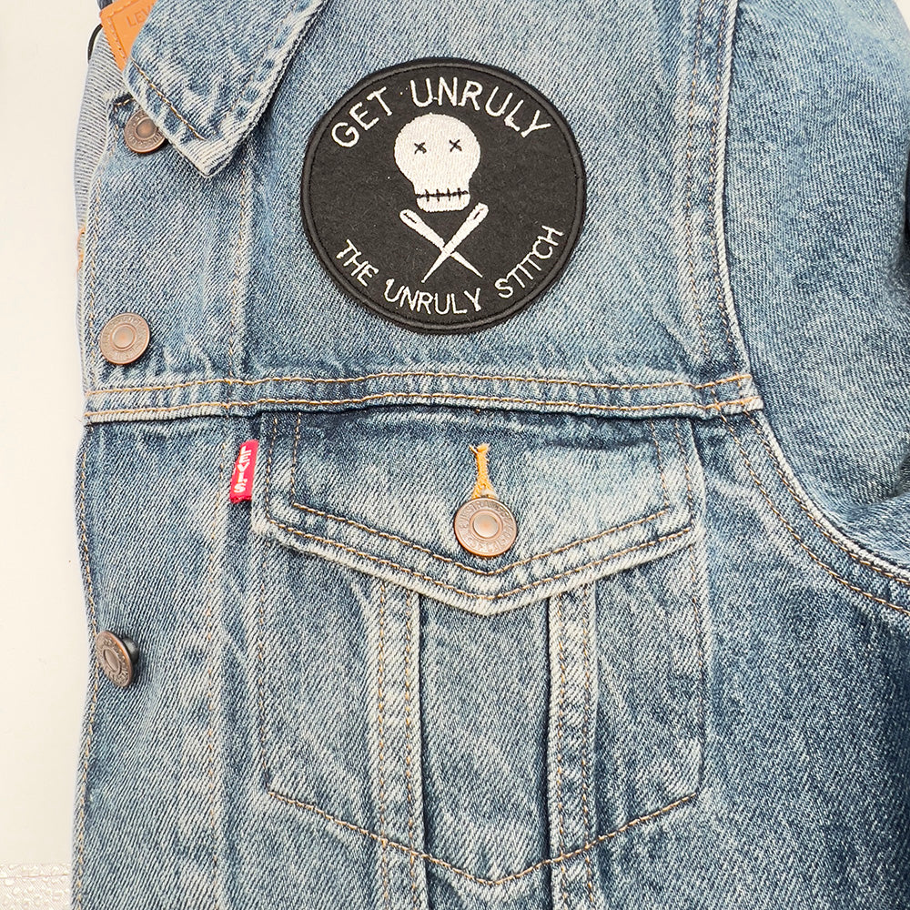 On denim jacket image: An embroidered felt patch with the text "Get Unruly The Unruly Stitch" surrounding a white skull and crossed needles is sewn onto the front pocket of a denim jacket. The patch is circular and features white embroidery on a black background.