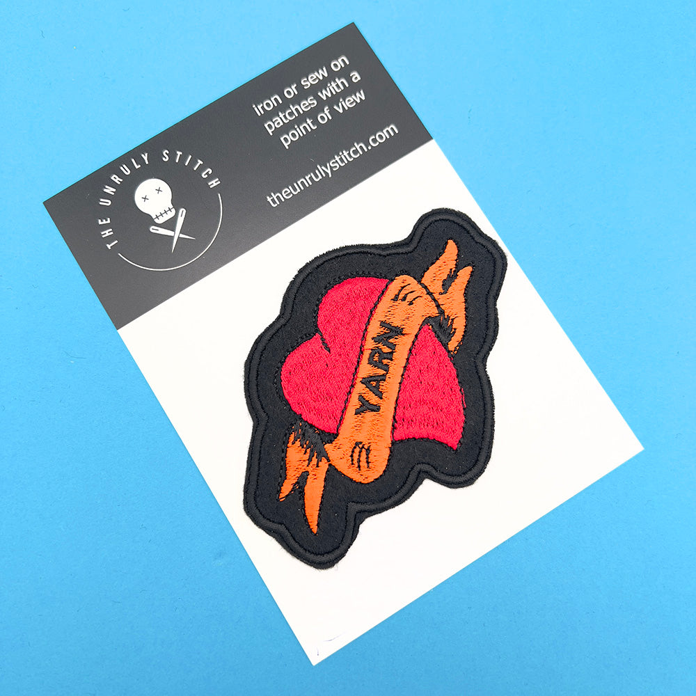 On card image: An embroidered felt patch shaped like a heart with a banner, displayed on a card. The patch features a red heart with an orange banner that reads "YARN," with black outlines and details.