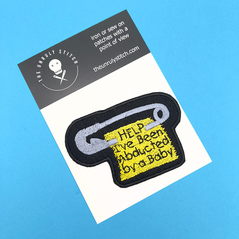 An embroidered felt patch with a safety pin and a yellow tag that reads "HELP I've Been Abducted by a Baby." The patch is mounted on a white card with the brand logo "The Unruly Stitch" at the top, all set against a blue background.