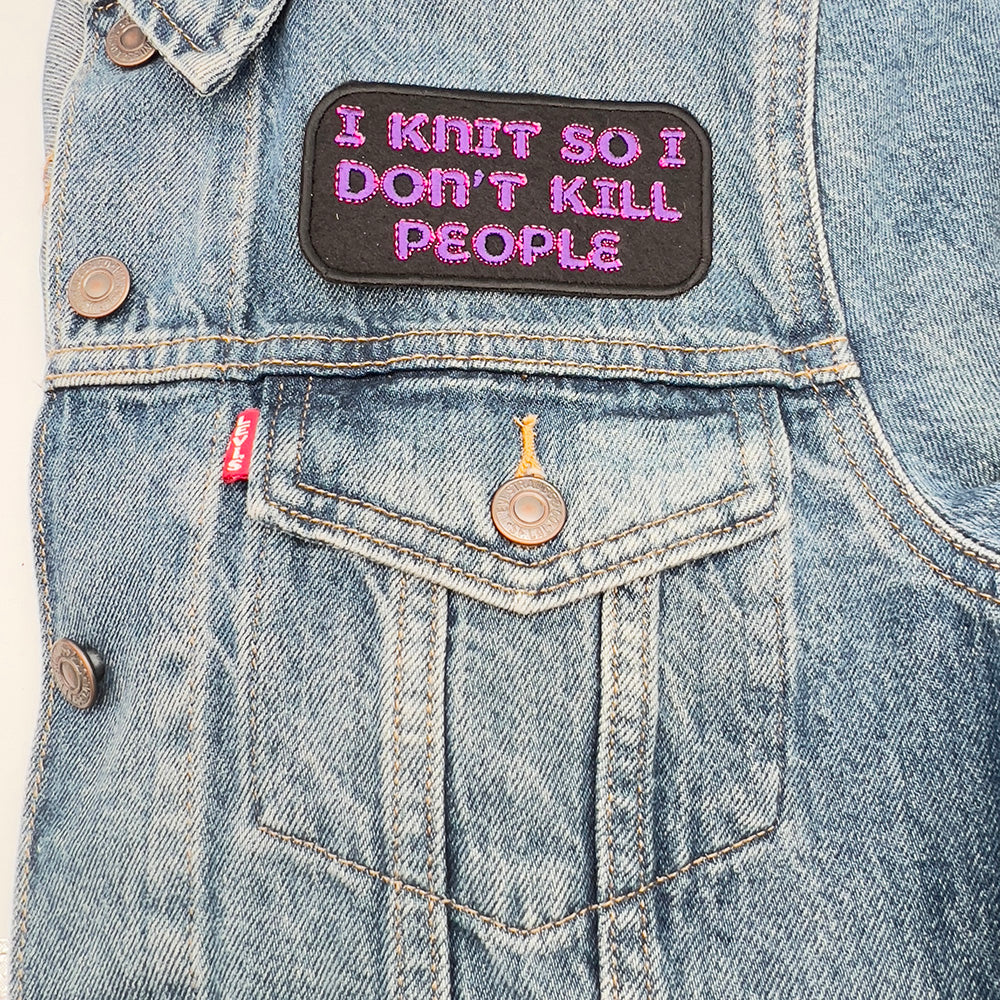 A denim jacket with an embroidered felt patch that reads "I KNIT SO I DON'T KILL PEOPLE" in pink and purple letters. The patch is sewn on the upper left side of the jacket, above the pocket.