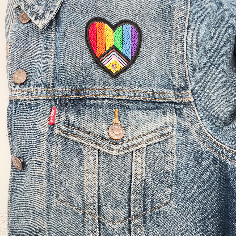 Heart-shaped Inclusive Pride flag embroidered patch attached to the pocket of a denim jacket.