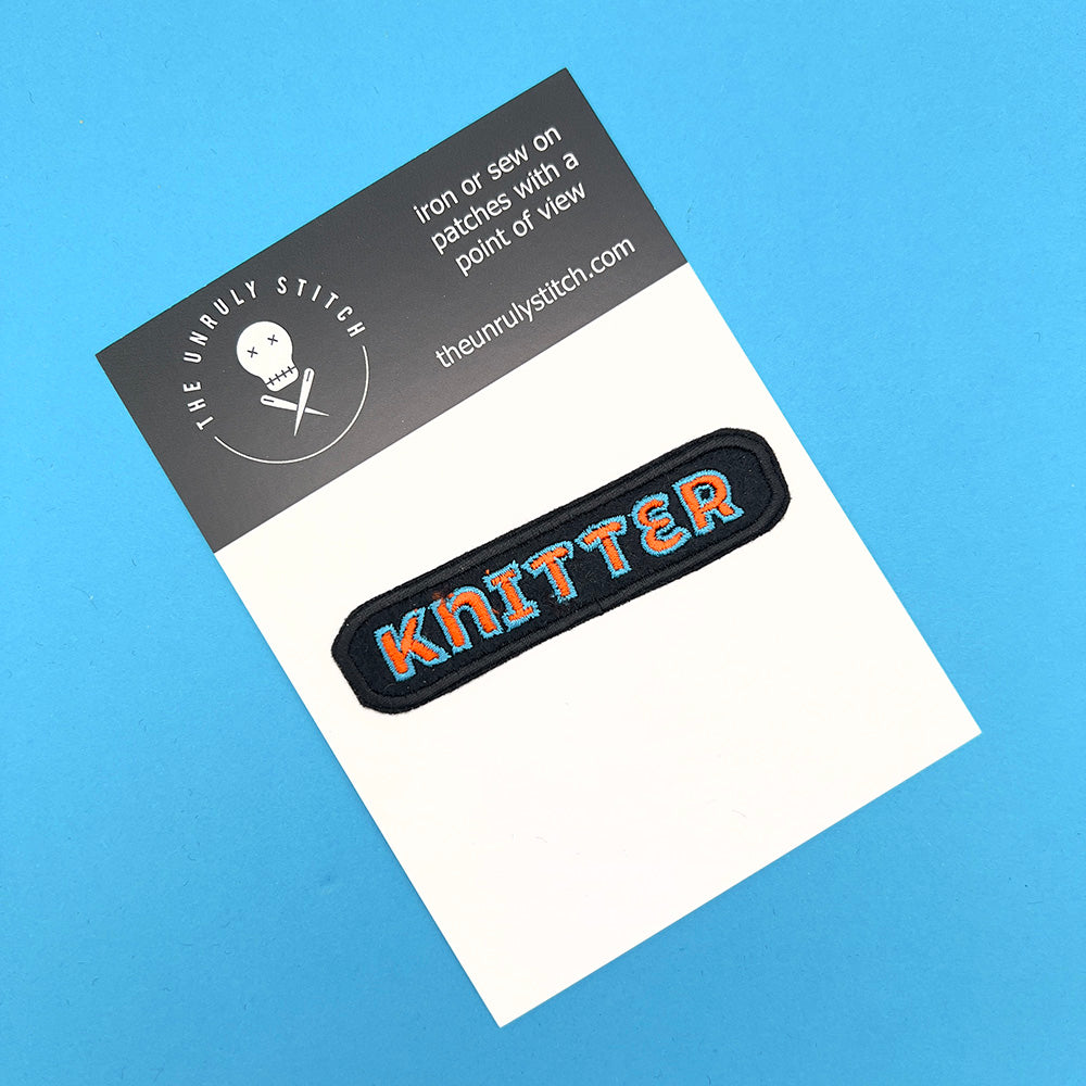 Embroidered felt patch with the word "Knitter" on a card. The patch features the word "Knitter" in orange letters outlined in light blue on a black background with a black border. The card has branding details and instructions for ironing or sewing on the patch.