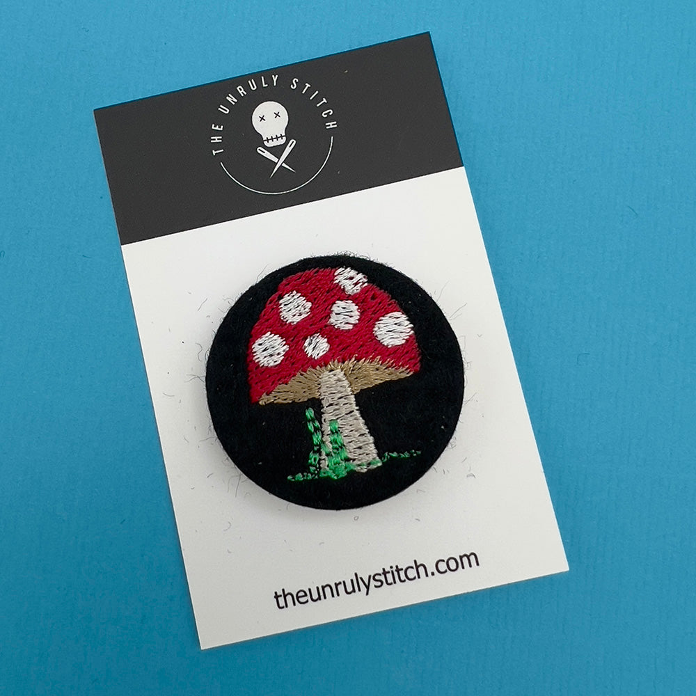 Embroidered felt badge depicting a red and white spotted mushroom on a black background, mounted on a branded card from The Unruly Stitch.