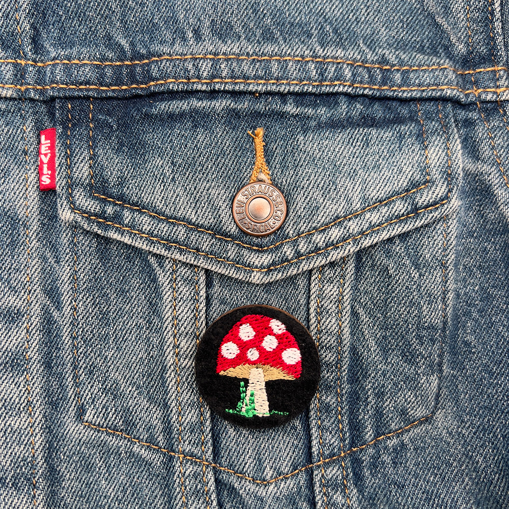 Embroidered felt badge with a red and white spotted mushroom design on a black background, pinned on the pocket of a denim jacket, designed by The Unruly Stitch