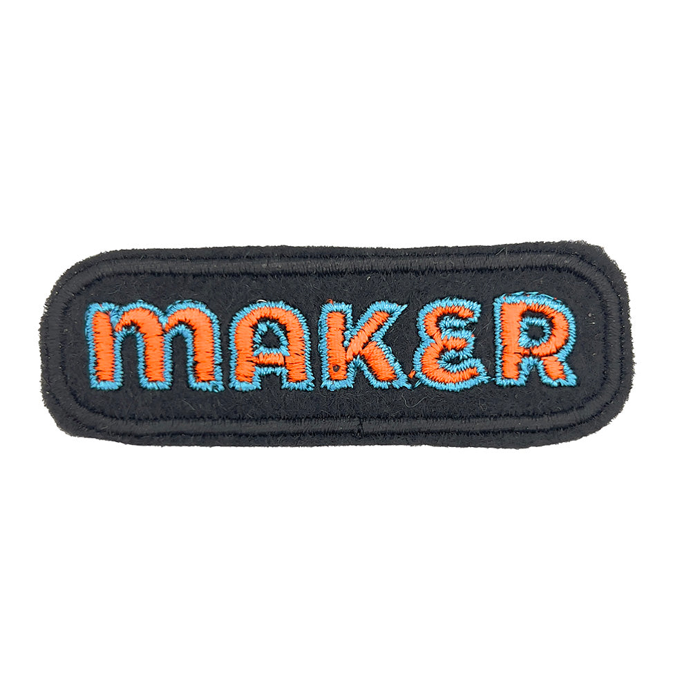 Close-up image: Close-up view of an embroidered felt patch with the word "MAKER" in orange letters outlined in blue, set against a black background with a rounded rectangular shape.
