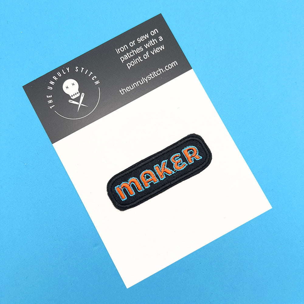 On card image: Embroidered felt patch with the word "MAKER" in orange letters outlined in blue, displayed on a white card with "The Unruly Stitch" branding, against a blue background.