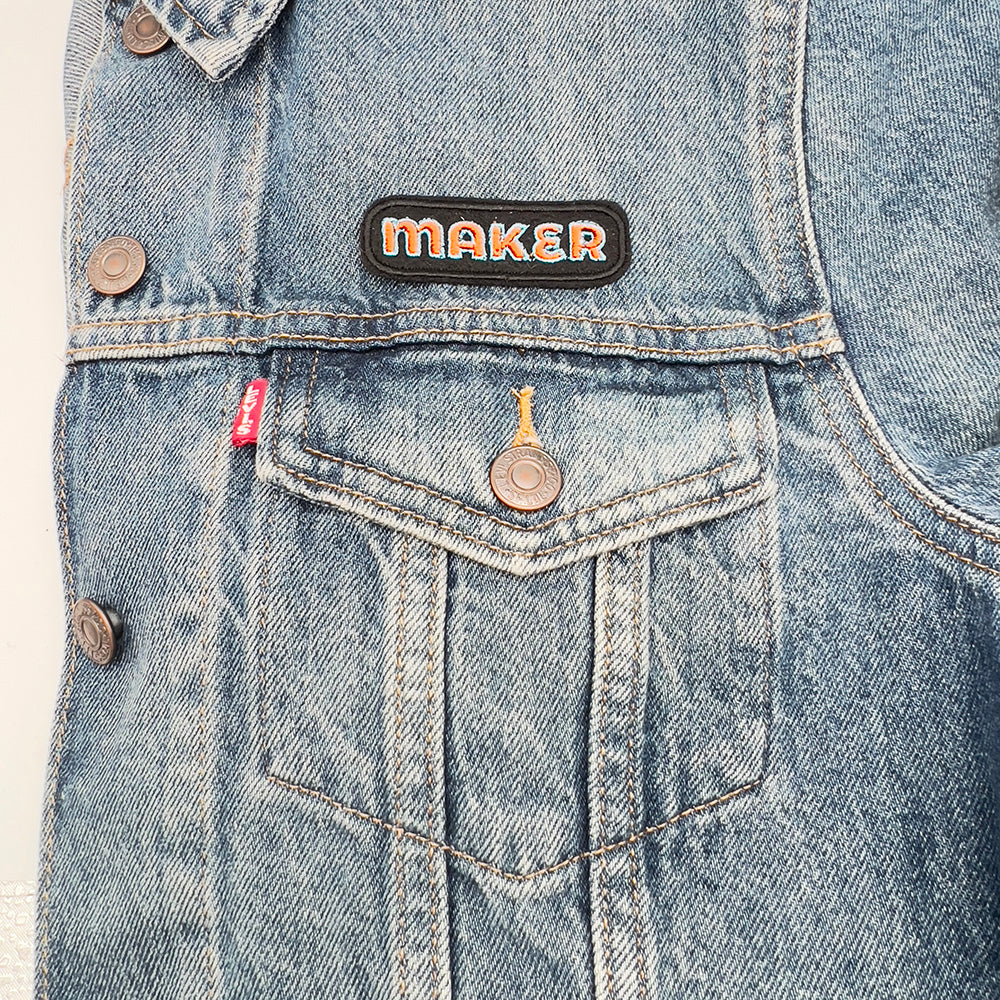 On denim jacket image: Embroidered felt patch with the word "MAKER" in orange letters outlined in blue, attached to the upper section of a denim jacket pocket, highlighting the contrast between the patch and the denim fabric.