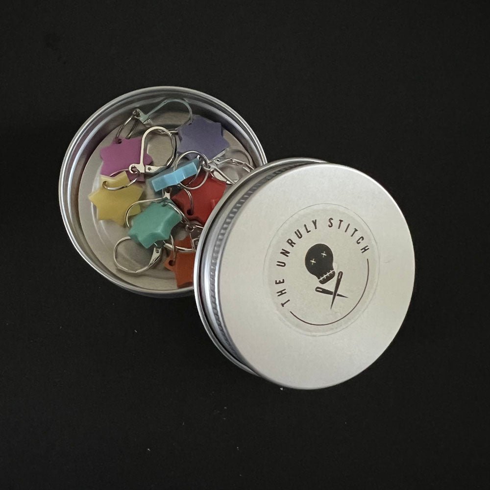 An open round metal tin with 'The Unruly Stitch' logo on the lid, containing various colorful stitch markers shaped like stars. The tin is placed on a black background.
