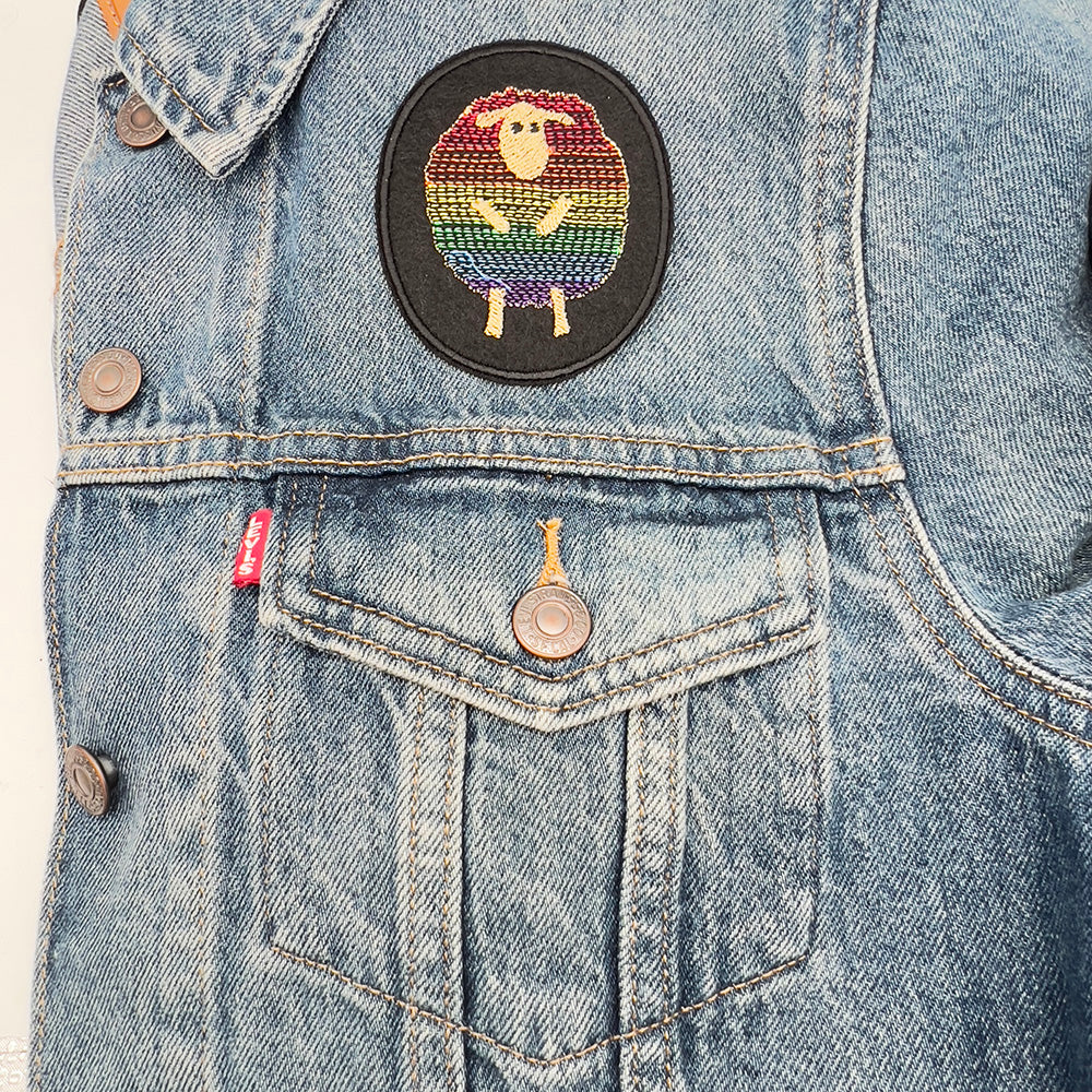 An embroidered felt patch of a sheep with a rainbow-colored body, outlined in beige, ironed onto a denim jacket just above the pocket. The patch stands out against the blue denim fabric.