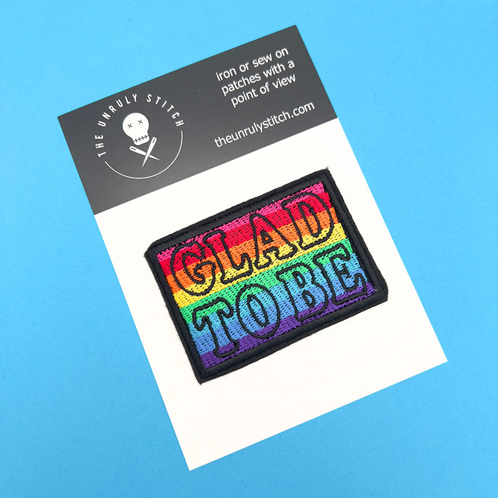 Embroidered felt patch with the words "GLAD TO BE" on a card. The patch has a rainbow gradient background with text outlined in black. The card features the branding "The Unruly Stitch" with a skull and crossed needles logo at the top.