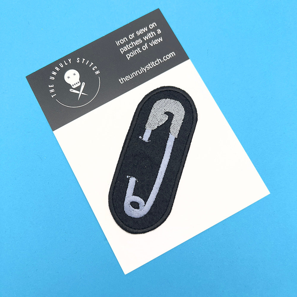 Embroidered felt patch with a metallic grey safety pin design on a black background, displayed on a branded card.