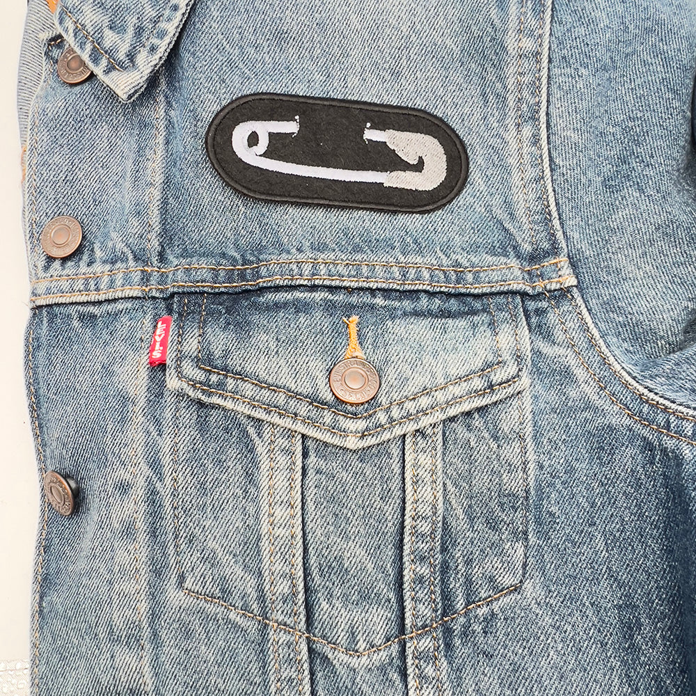 Image on Denim Jacket: Embroidered felt patch with a white safety pin design on a black background, sewn onto the upper left side of a light blue denim jacket.
