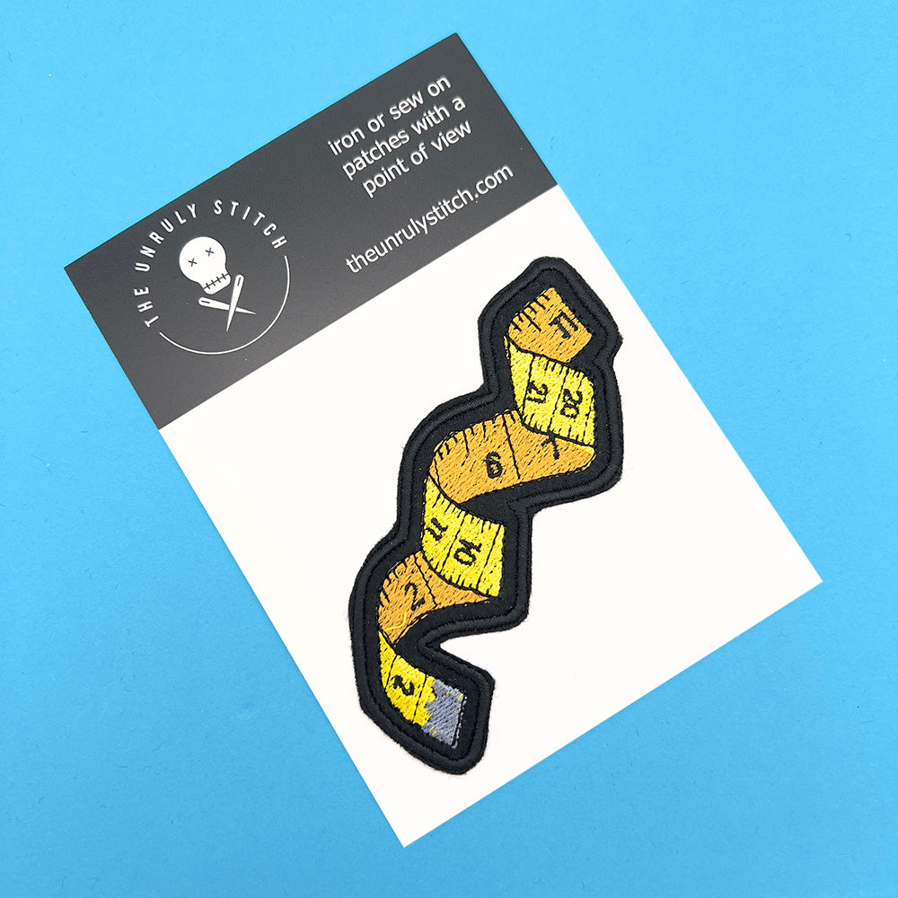 An embroidered felt patch shaped like a winding tape measure, displayed on a white card with "The Unruly Stitch" branding at the top. The patch features a tape measure with numbers ranging from 1 to 12, depicted in yellow and orange sections with black outlines and details. The card sits on a blue background.