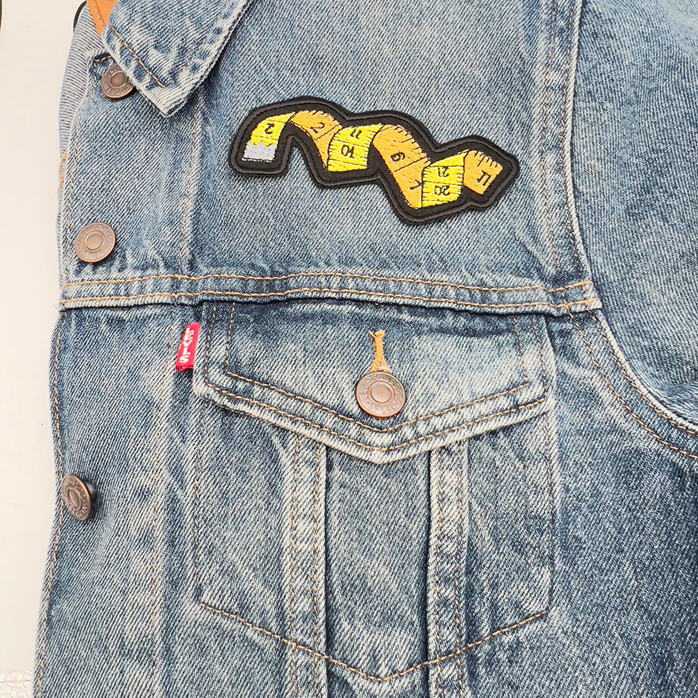 An embroidered felt patch shaped like a winding tape measure, ironed above the pocket of a blue denim jacket. The patch features a tape measure with numbers ranging from 1 to 12, depicted in yellow and orange sections with black outlines and details