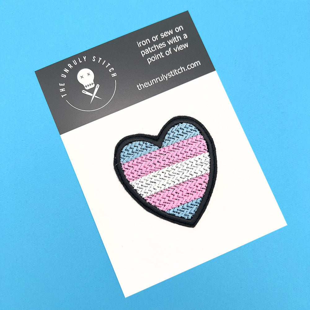 A heart-shaped Transgender Pride flag patch with light blue, pink, and white horizontal stripes, attached to a white card. The card sits on a blue background and includes branding details of "The Unruly Stitch."