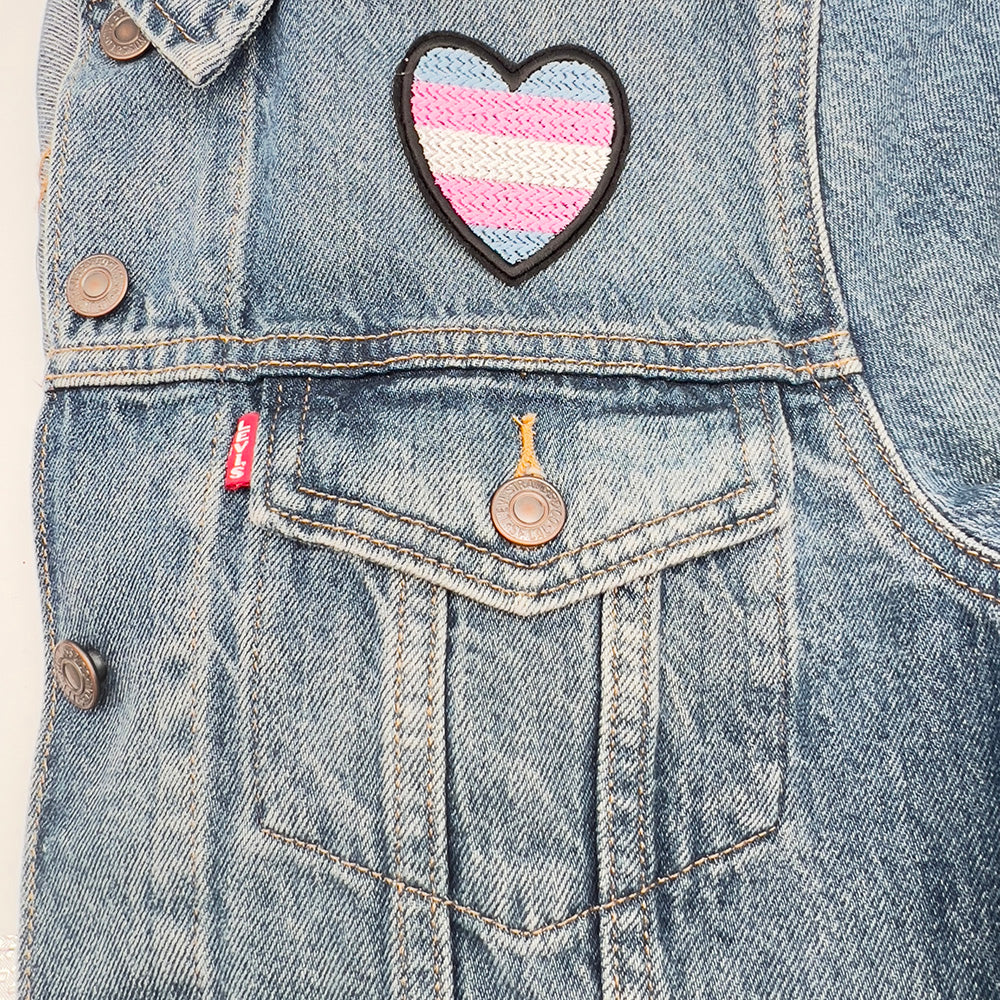 A heart-shaped patch with the Transgender Pride flag colors (light blue, pink, and white horizontal stripes) ironed above the pocket of a denim jacket, displaying the patch prominently on the upper section of the jacket.
