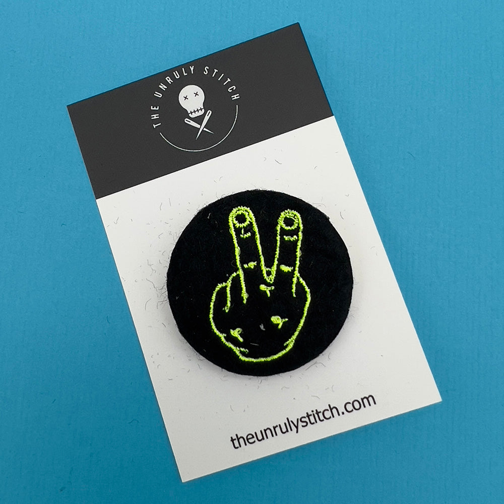 Branded card with felt badge with an embroidered hand with two fingers up in yellow threads on a black background, designed by The Unruly Stitch.