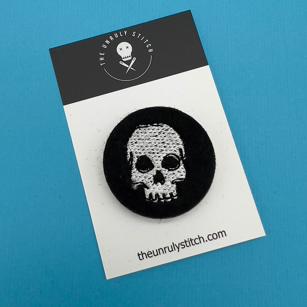 A black felt badge with a white embroidered skull displayed on a branded card. The card has 'The Unruly Stitch' logo and website URL at the bottom.