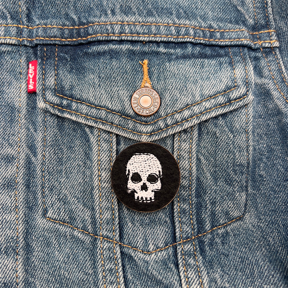 A black felt badge with a white embroidered skull attached to the front pocket of a blue denim jacket. The badge contrasts sharply with the denim fabric