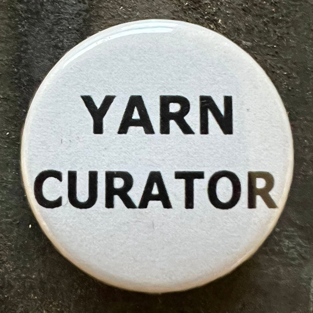 Close-up of a white YARN CURATOR pin badge with black text.