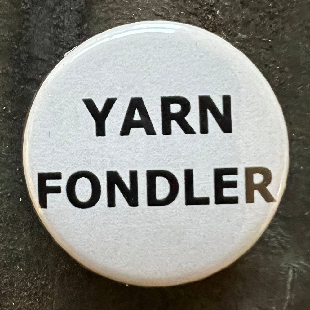 Close-up of a white YARN FONDLER pin badge with black text.