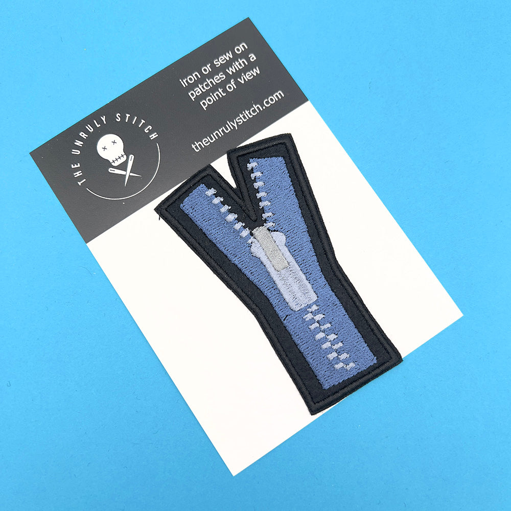 An embroidered felt patch shaped like an uppercase "Y" displayed on a card. The patch features a blue zipper with white teeth and a white pull tab, depicted with black outlines and details. The card background is light blue with branding information at the top.