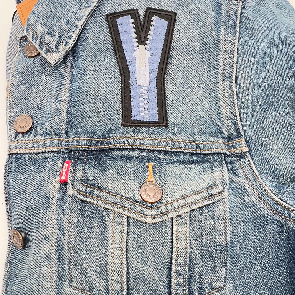 An embroidered felt patch shaped like an uppercase "Y" attached to the front of a denim jacket. The patch features a blue zipper with white teeth and a white pull tab, depicted with black outlines and details. The jacket shows buttons and a visible pocket flap.