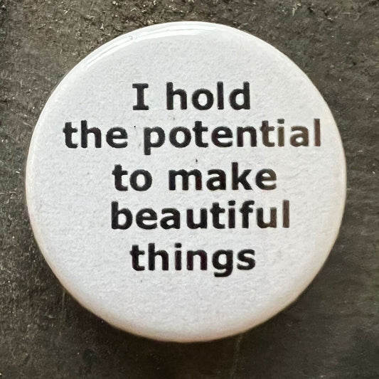 Close-up of a white I hold the potential to make beautiful things pin badge with black text.