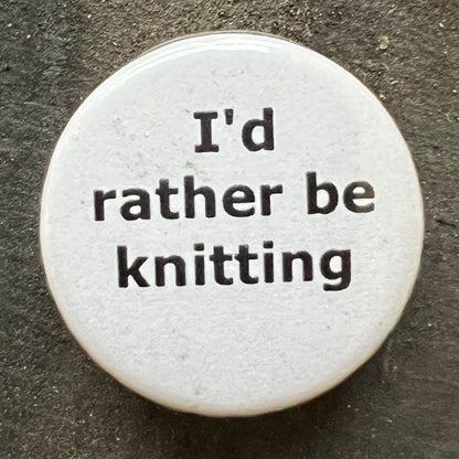 Close-up of a white I'd rather be knitting pin badge with black text.