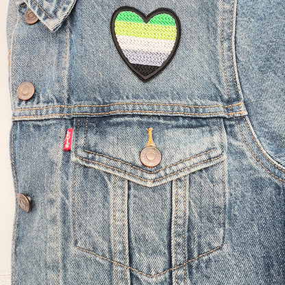 Aromantic Pride flag heart-shaped embroidered felt patch attached to the front pocket of a blue denim jacket from The Unruly Stitch