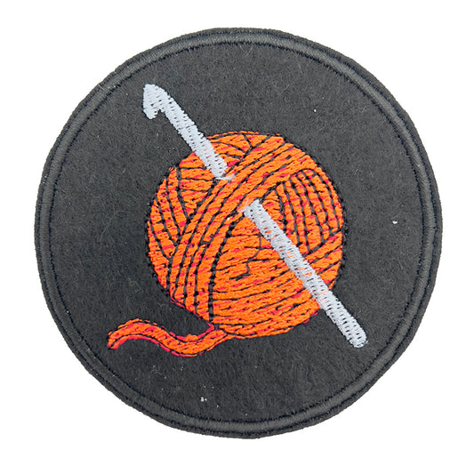 Close-up view of an embroidered felt patch depicting a ball of orange yarn with a silver-coloured crochet hook inserted through it. The patch is circular with a black background and detailed stitching.