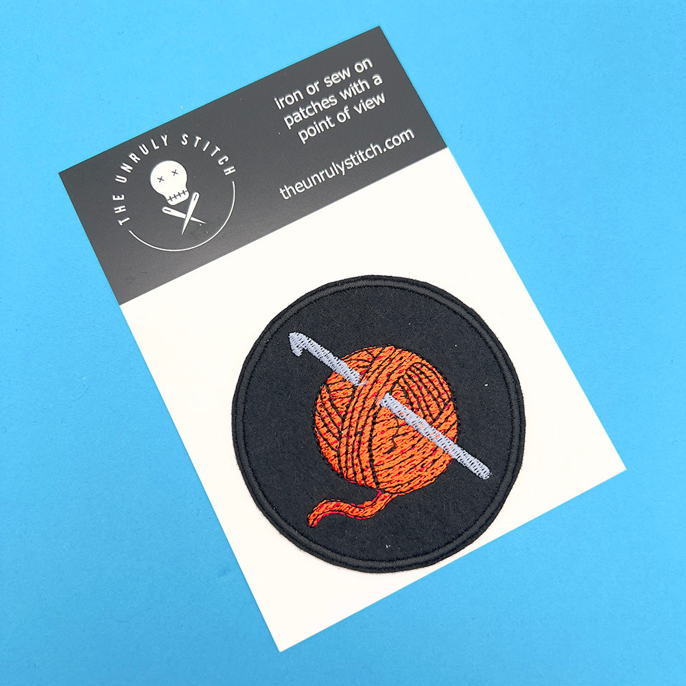 Image of an embroidered felt patch depicting a ball of orange yarn with a silver-coloured crochet hook inserted through it, displayed on a card with branding. The patch is circular with a black background and detailed stitching.