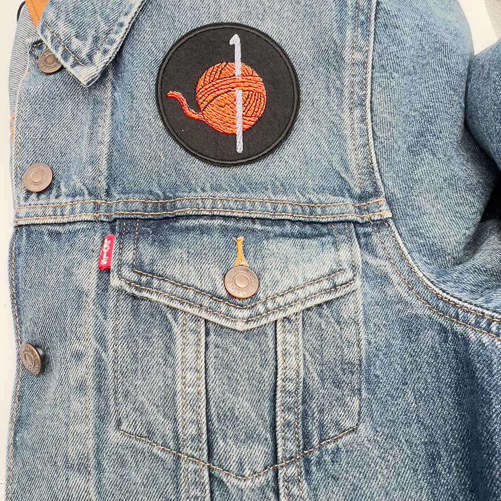 Embroidered felt patch depicting a ball of orange yarn with a silver-coloured crochet hook inserted through it, sewn onto the pocket area of a blue denim jacket. The patch is circular with a black background and detailed stitching.