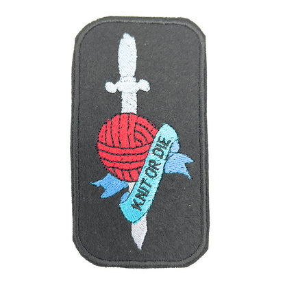 Close-up view of an embroidered felt patch depicting a red ball of yarn with a silver dagger through it. A blue banner with the words "Knit or Die" wraps around the yarn and dagger.
