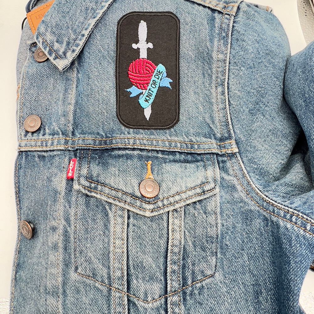 The embroidered felt patch featuring a red ball of yarn with a silver dagger through it and a blue banner with the words "Knit or Die" is sewn onto the front pocket of a denim jacket.