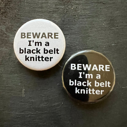 A close-up image of two round pin badges on a dark surface. One badge is white with black text that reads "BEWARE I'm a black belt knitter," and the other badge is black with the same text in white.  