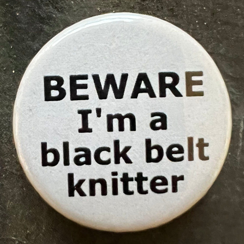 A close-up image of a single white round pin badge with black text that reads "BEWARE I'm a black belt knitter." The badge is placed on a dark background.