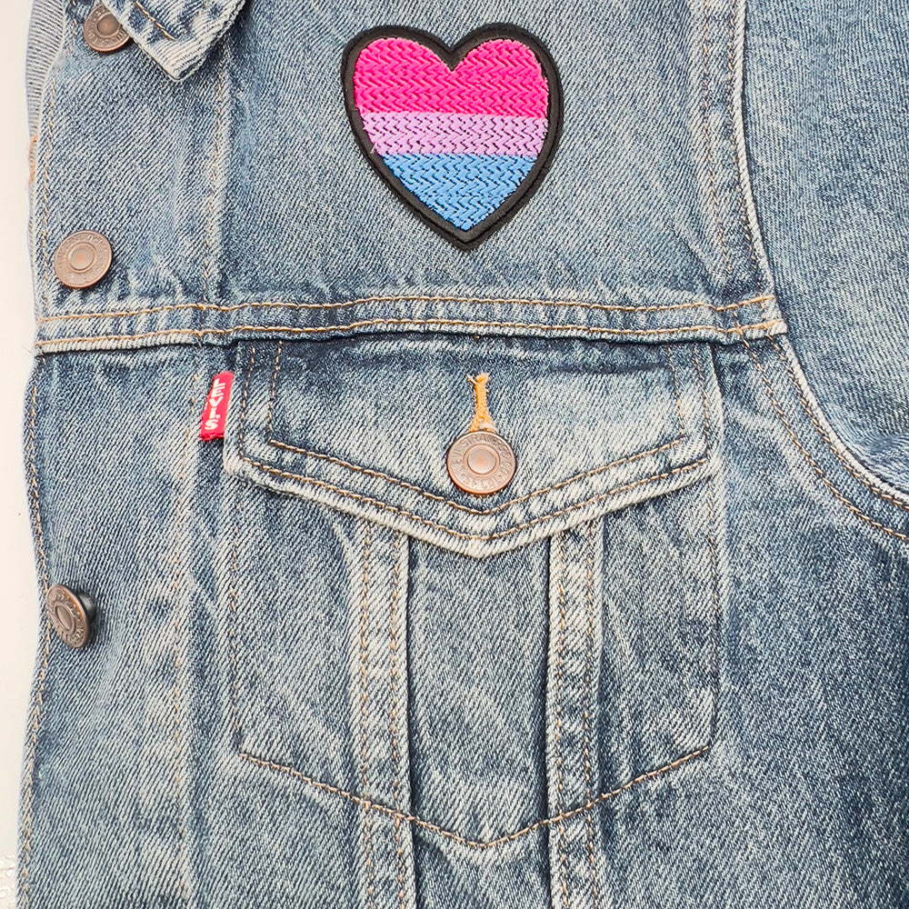 Bisexual pride flag heart-shaped embroidered felt patch attached to the front pocket of a denim jacket. The patch displays horizontal stripes in pink, purple, and blue.