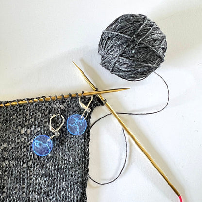 Close-up of a black knitted fabric on a gold knitting needle, with a ball of gray yarn beside it. Two blue circular perspex stitch markers with constellation designs are clipped onto the knitted fabric.