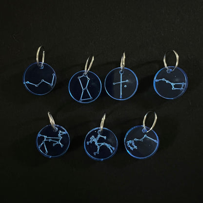 Seven blue circular perspex stitch markers with various constellation designs on silver jump rings, arranged on a black background.