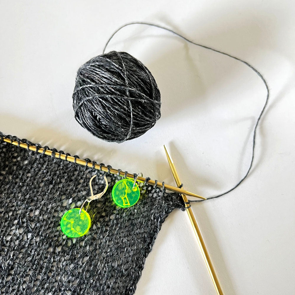 Close-up of a black knitted fabric on a gold knitting needle, with a ball of gray yarn beside it. Two neon green circular perspex stitch markers with constellation designs are clipped onto the knitted fabric.