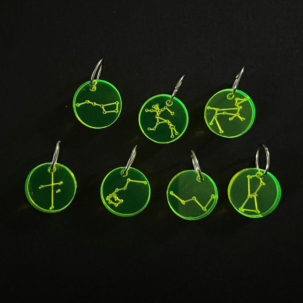 Seven neon green circular perspex stitch markers with various constellation designs on silver jump rings, arranged on a black background.