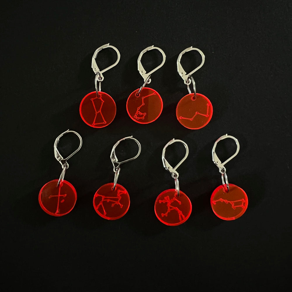 Seven red circular perspex stitch markers with various constellation designs, attached to leverback hooks, arranged on a black background.